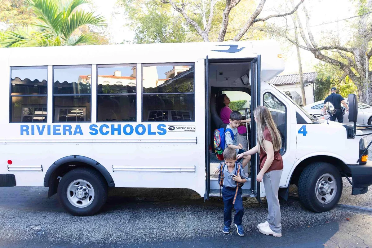 Students exiting white Riviera Schools bus while staff member assists with arrival at tree-lined campus drop-off area
