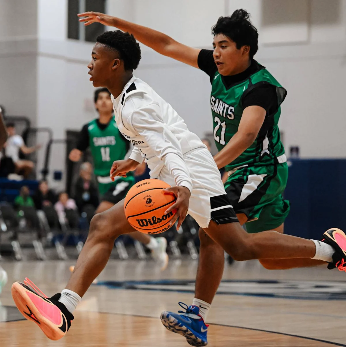 Two basketball players in action, one in white jersey dribbling while defender in green Saints #21 jersey guards with raised arm