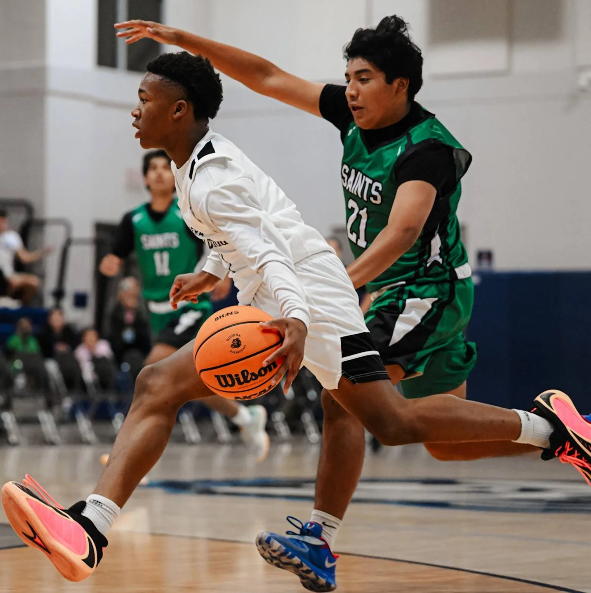 Two basketball players in action, one in white jersey dribbling while defender in green Saints #21 jersey guards with raised arm