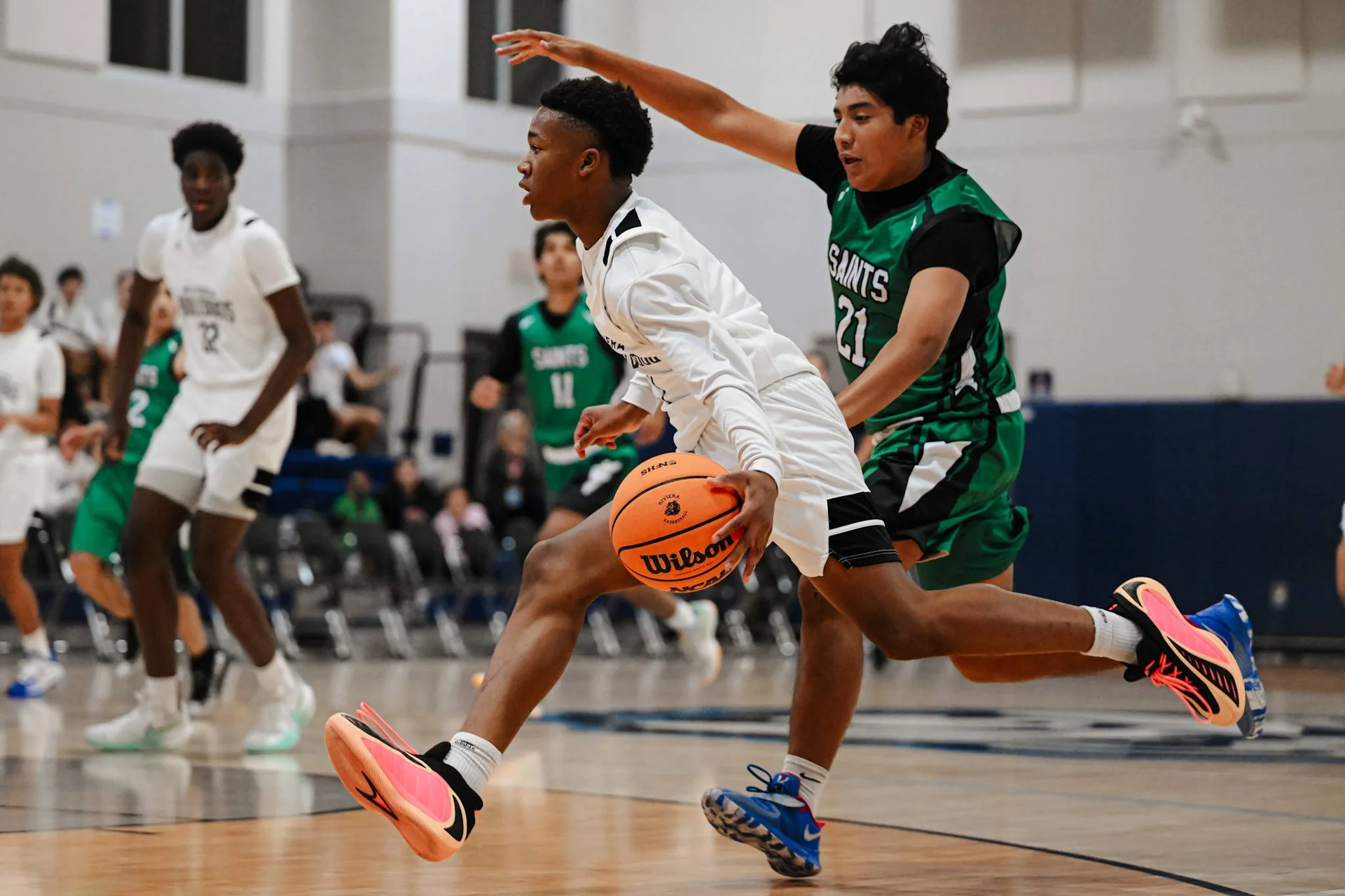 Two basketball players in action, one in white jersey dribbling while defender in green Saints #21 jersey guards with raised arm