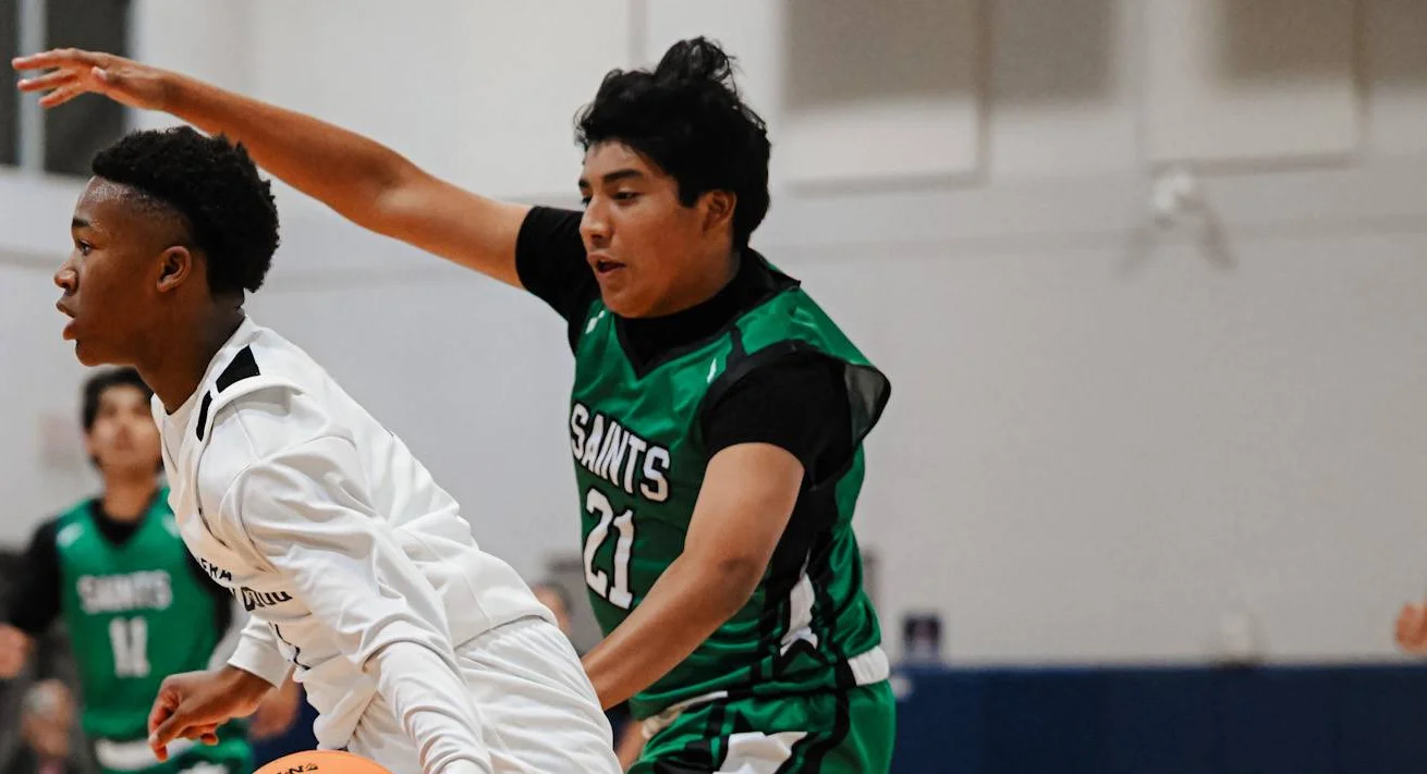 Two basketball players in action, one in white jersey dribbling while defender in green Saints #21 jersey guards with raised arm