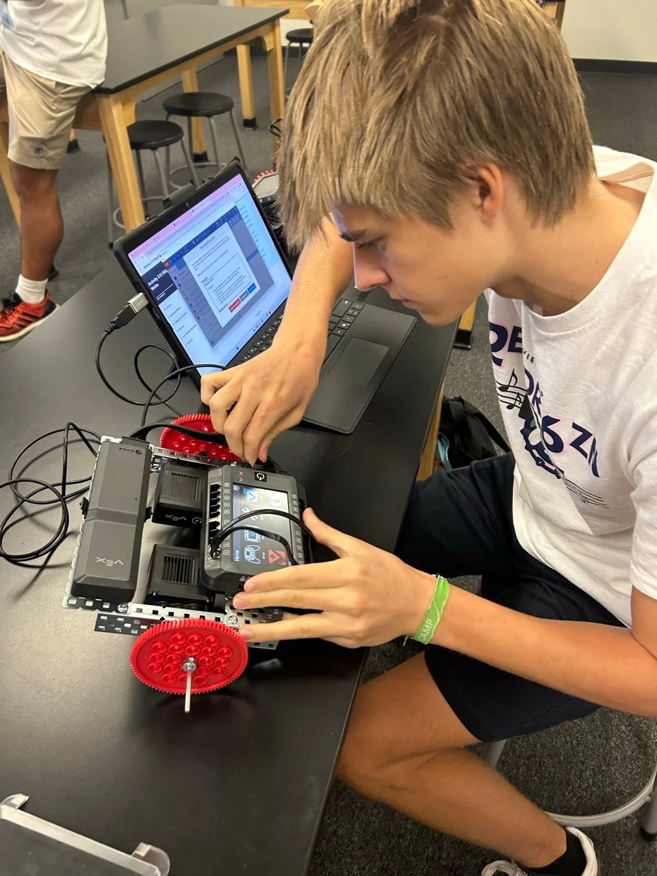 Student working on a VEX robotics project with red gear wheels, programming on a laptop in a classroom setting