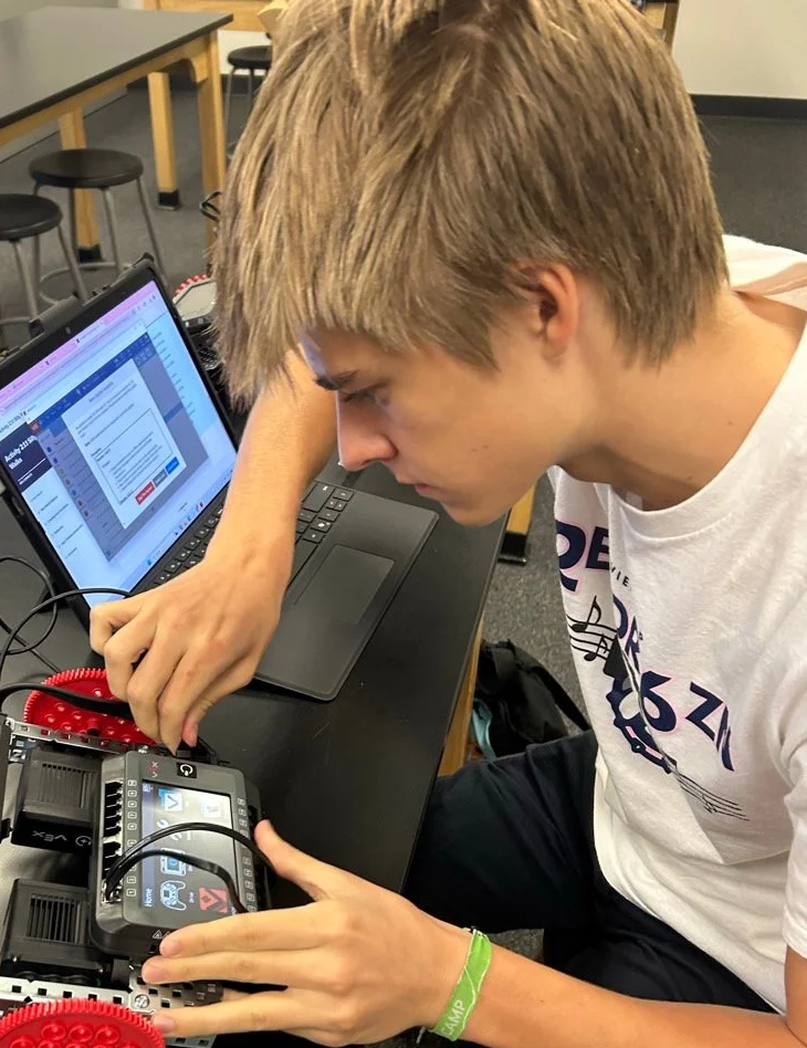 Student working on a VEX robotics project with red gear wheels, programming on a laptop in a classroom setting